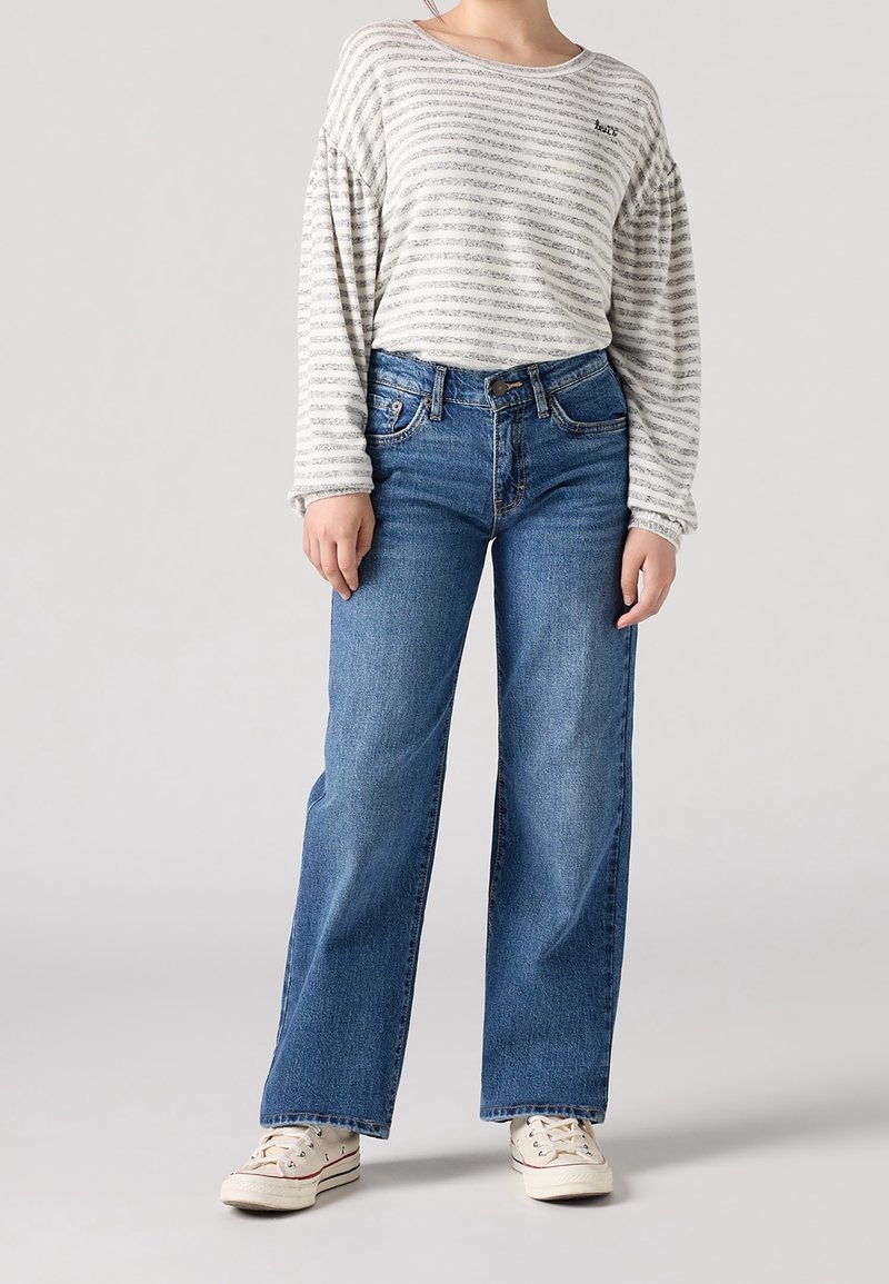 Person som bär en ljusgrå randig långärmad skjorta, blå vida jeans och vita sneakers som står framför en ljus bakgrund.