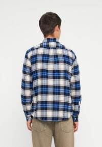 Camisa de flanela de xadrez azul e branca com colarinho abotoado e mangas longas. Apresenta um acabamento arredondado e uma textura suave.