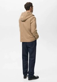 Beige hættejakke med tætsiddende design, vist bagfra, kombineret med mørke denimjeans og sorte sko, på en ensfarvet hvid baggrund.