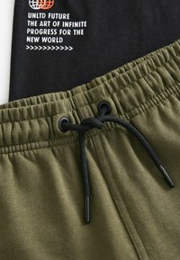 Olivenfarvede sweatpants med en sort elastisk talje, der har snøre og trykt tekst i hvid og rød på det sorte stof ovenfor.