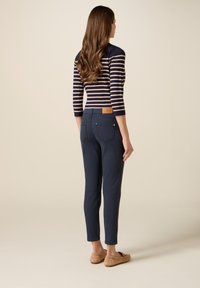 Vrouw met lang bruin haar, gekleed in een marineblauw gestreept shirt, donkerblauwe strakke broek en beige mocassins, staand tegen een eenvoudige beige achtergrond.