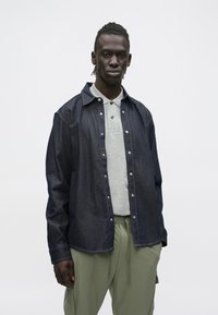 Młody mężczyzna o ciemnej skórze i plecionych włosach ma na sobie ciemną dżinsową koszulę na szarej koszulce polo oraz oliwkowo-zielone spodnie na jednolitym tle.