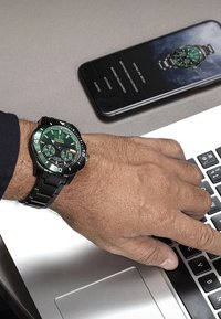 Montre noire avec un cadran vert et des marqueurs luminescents, dotée d'un bracelet en acier inoxydable, présentée aux côtés d'un smartphone et d'un ordinateur portable.