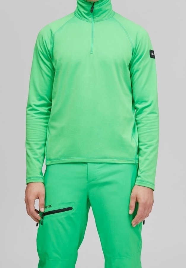 Homme portant un haut vert vif à manches longues avec col zippé et un pantalon vert assorti avec une poche zippée noire sur la cuisse, sur fond gris.