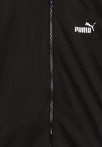 Črna jakna na zadrgo iz gladkega blaga, ki ima bel Puma logotip na prsih in tanko zadrgo z teksturiranim zategom.