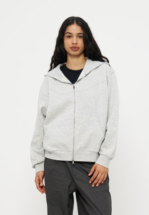 Jeune femme aux longs cheveux noirs portant un sweat à capuche gris clair zippé et un pantalon foncé, debout devant un fond blanc uni.