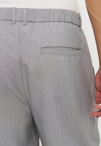 Vue arrière d'un pantalon gris clair avec une ceinture élastique et une unique poche passepoilée à l'arrière, porté par une personne portant une chemise blanche.