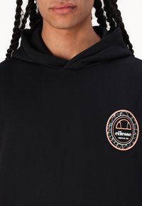 Svart hettegenser med kengurulomme, utstyrt med et rundt ellesse-logo i hvitt og rosa på brystet, laget av mykt bomullsmateriale.
