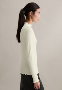 Kremowy, długi rękaw top o prążkowanej fakturze, z wysokim dekoltem i falistymi krawędziami; w połączeniu z ciemnymi spodniami, widoczny z boku.