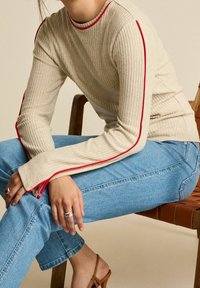 Beżowa, prążkowana bluzka z długim rękawem i czerwonymi akcentami, w połączeniu z jasnoniebieskimi dżinsami. Modelka siedzi na drewnianym krześle.