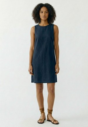 Femme aux cheveux bouclés portant une robe bleu foncé sans manches longueur genou et des sandales plates marron à brides, debout devant un fond blanc uni.