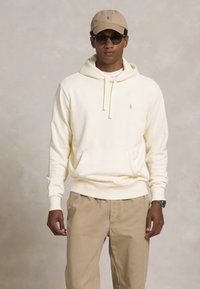 Crème-kleurige hoodiesweater met een voorzak en een trekkoord. Gepaarde met beige broek en een bijpassende pet. Donkere zonnebril.
