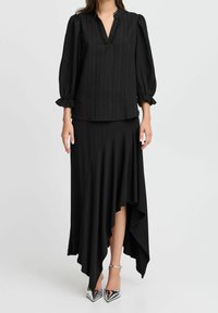 Blouse noire texturée avec un col en V et des manches bouffantes, associée à une jupe noire asymétrique avec un ourlet à volants, accompagnée de talons argentés.