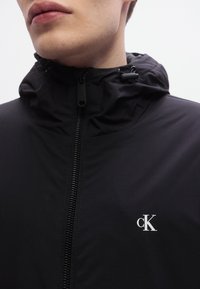 Μαύρο μπουφάν με φερμουάρ και κουκούλα, φτιαγμένο από υφή υφάσματος. Διαθέτει μικρό λευκό λογότυπο "ck" στο στήθος και λείο φερμουάρ.