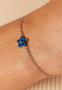 Zilveren kettingarmband met een blauw steenvormige bloemontwerp met zes bloemblaadjes en een centrale steen, geplaatst tegen een huidtextuur.