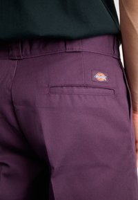Närbild av vinröda Dickies-byxor med bakficka och synlig varumärkeslapp ovanför fickan, tillsammans med en svart topp.