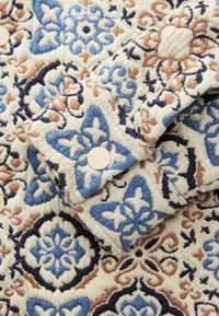 Tekstil med blomsterprint i blå, creme og rust, der har et blødt, struktureret materiale og runde knapper. Udstyret med detaljerede og indviklede mønstre.