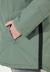 Grön jacka med matta ytor, med en sidoficka och svarta detaljer längs sömmen. Slät textur, något oversized passform.