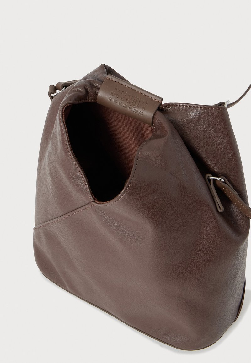Sac à bandoulière en cuir marron avec ouverture supérieure, intérieur en tissu visible et étiquette en cuir cousue portant un logo embossé numéroté.