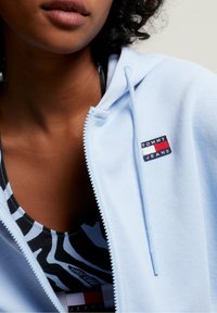 Mujer vistiendo una sudadera ligera azul de Tommy Jeans parcialmente desabrochada sobre una camisilla de estampado de cebra en blanco y negro con el logo de Tommy Jeans.