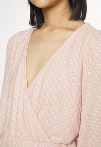 Vestido envolvente rosa claro com tecido texturizado em padrão de colmeia, decote em V profundo e mangas longas e esvoaçantes.