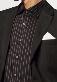 Abito nero con una camicia a righe nere che presenta sottili strisce verticali bordeaux e un fazzoletto bianco nel taschino. Colletto abbottonato con bottoni in contrasto.