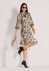 Frau in einem satinartigen Leopardenmuster-Kleid mit Schleifenkragen, schwarzen Peep-Toe-Ankelboots, großen Sonnenbrillen, goldener Schmuck und einer rosa Handtasche.