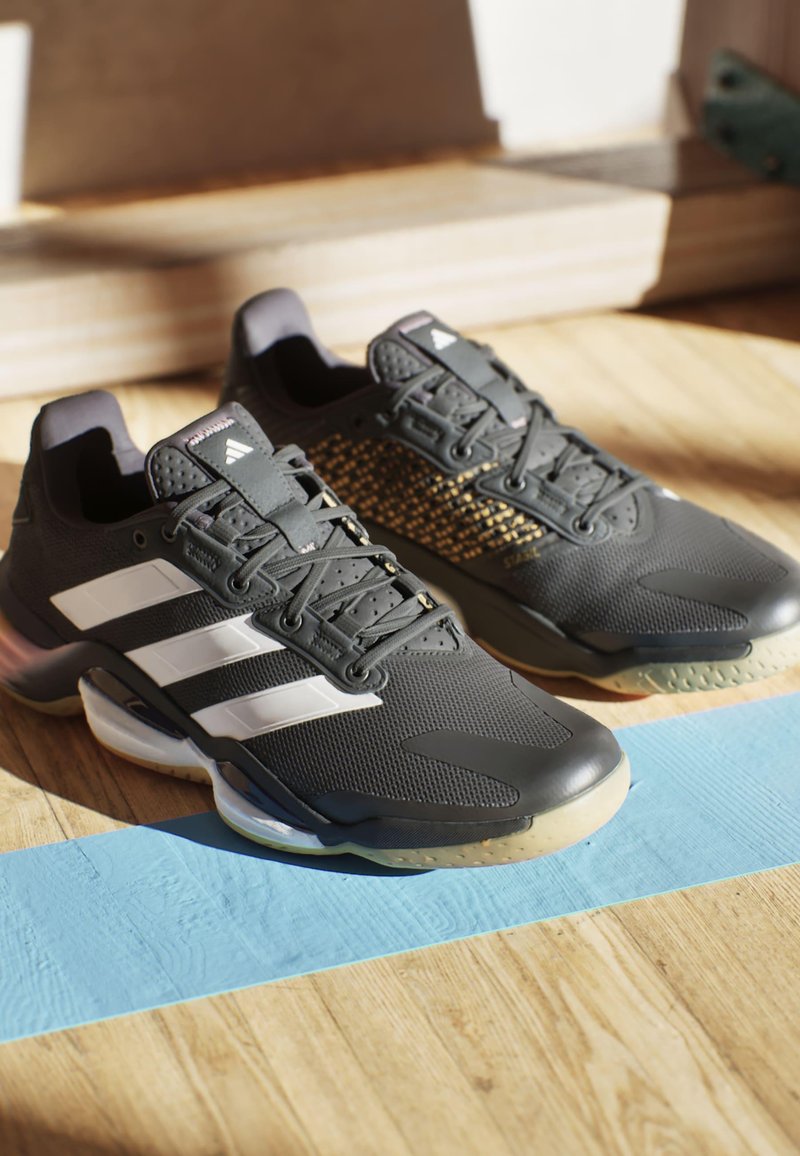 Scarpe da ginnastica nere con superficie testurizzata, caratterizzate da strisce bianche e dettagli dorati, punta rotonda e suole in gomma trasparente.