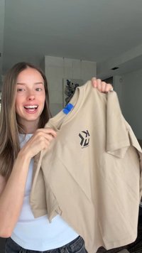 Young woman with long brown hair holds up a beige t-shirt with a black logo near the neckline, smiling and speaking indoors.