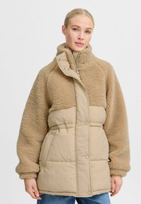 Beige polstret vinterjakke med en fuzzy, tekstureret overdel, høj krave og trykknap-/knaplukninger. Har oversized ærmer og sidelommer.