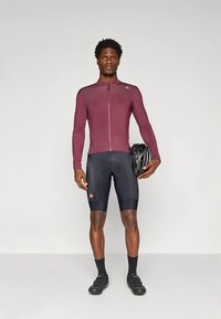 Bordó hosszú ujjú kerékpáros mez cipzárral; fekete kerékpáros rövidnadrág; fekete zokni; fekete kerékpáros cipő; fekete sisakot tartva.