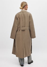 Dlhý béžový trenčkot s opaskom v oblasti pása, širokými rukávmi a rovnou siluetou. Má hladkú, matnú textúru.