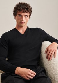 Schwarzer V-Ausschnitt-Pullover mit langen Ärmeln, aus weichem Material, mit gerippten Bündchen und figurnahem Schnitt, kombiniert mit dunklen Hosen.