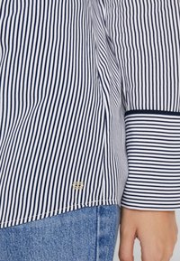 Camicia a righe in navy e bianco, con maniche lunghe, design dritto e un accento in logo dorato vicino all'orlo. Il tessuto appare liscio.