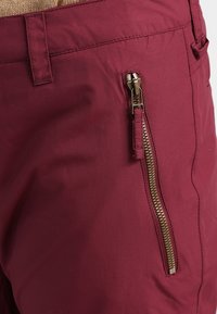 Bermuda merah marun dengan ritsleting samping dan perlengkapan emas, terbuat dari kain halus dan ringan dengan potongan yang bersih dan terstruktur.