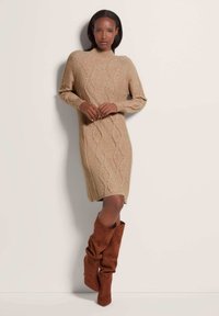 Beige gebreide jurk met kabelpatronen, met een hoge halslijn en lange mouwen. In combinatie met bruine slouchy kniehoge laarzen.