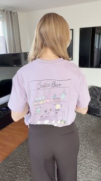 Persona con cabello rubio hasta los hombros lleva una camiseta lavanda con una ilustración colorida de "Juice Bar" en la espalda, de pie en el interior.