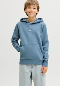 Χαμογελαστό αγόρι που φοράει μπλε φούτερ "Jack & Jones" με τα χέρια μερικώς στην μπροστινή τσέπη, συνδυασμένο με ανοιχτόχρωμο τζιν, σε απλό φόντο.