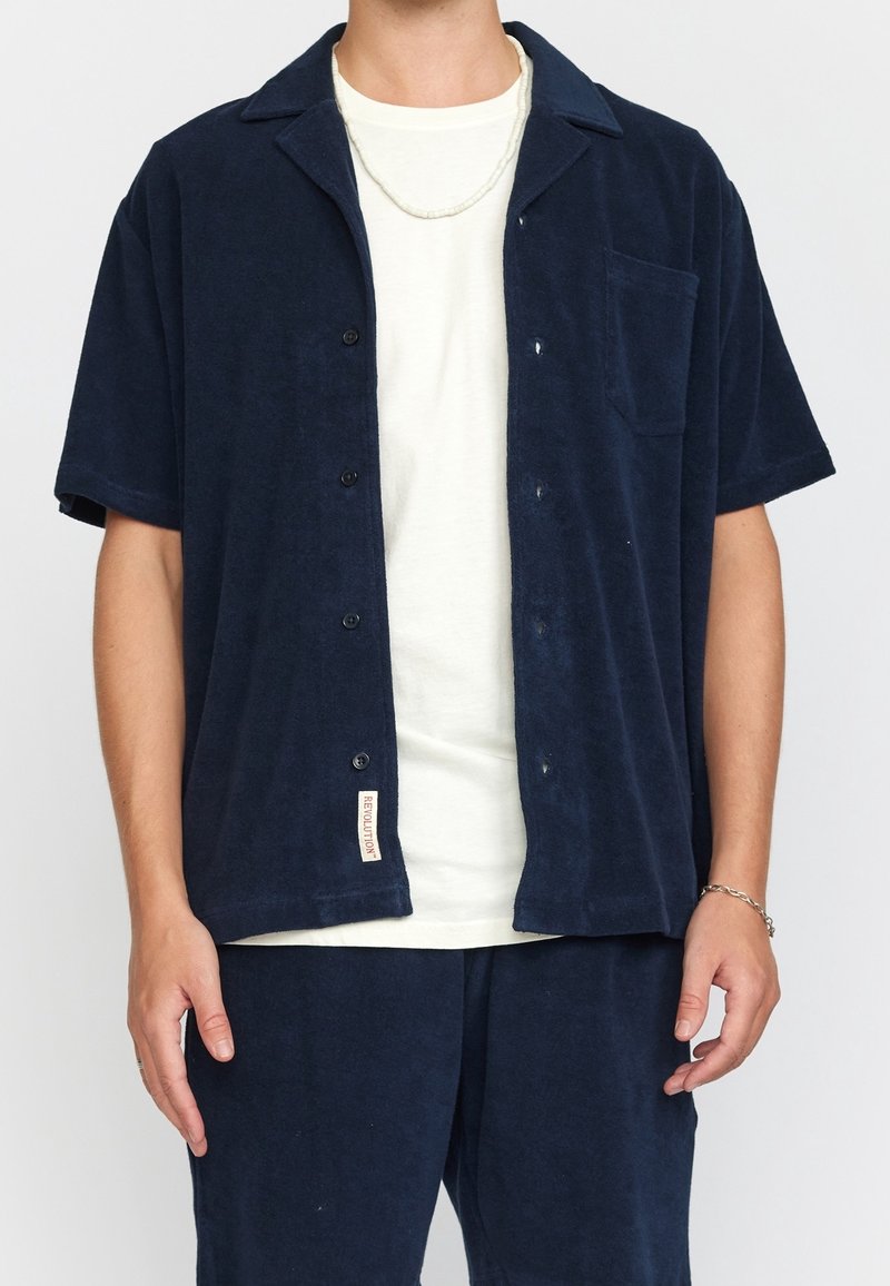 Personne portant une chemise bleu marine à manches courtes ouverte sur un t-shirt blanc avec un pantalon bleu marine assorti et un bracelet chaîne au poignet droit.