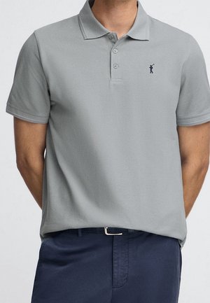 Hombre vistiendo una camisa polo de manga corta color gris claro con un pequeño logo oscuro en el pecho izquierdo y pantalones azul oscuro con un cinturón tejido negro.