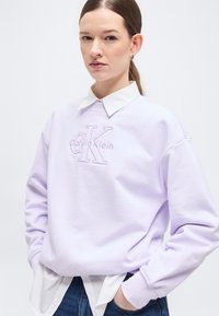 Νεαρή γυναίκα με καστανά μαλλιά που είναι μαζεμένα πίσω φοράει ένα ανοιχτό μωβ φούτερ Calvin Klein πάνω από ένα λευκό πουκάμισο με γιακά και μπλε τζιν, με τα χέρια στις τσέπες.