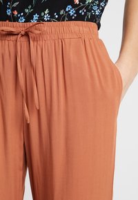 Pantalon à taille élastique froncée de couleur rouille avec un cordon de serrage. Le tissu semble léger et lisse, avec des poches latérales visibles.