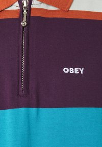 Polo triko se zipem, s vodorovnými pruhy v purpurové, tyrkysové a oranžové barvě, na hrudi bílá vyšitá logo "OBEY".