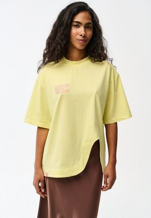 Kobieta z długimi kręconymi włosami nosi luźny żółty T-shirt z małym napisem i brązową spódnicę, stojąc na jednolitym białym tle.