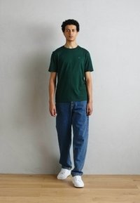 Zielona koszulka z krótkim rękawem, luźne niebieskie dżinsy i białe sneakersy; stoi na tle jasnoszarym, wyraźna tekstura naturalna.