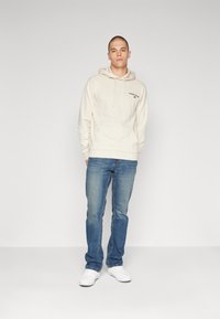 Felpa beige con tasca frontale e logo, abbinata a jeans in denim blu e sneakers bianche su uno sfondo neutro.