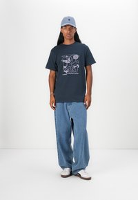 Navy t-shirt with abstract pink graphic, paired with light blue baggy jeans and white sneakers. Accessorized with a light blue cap.