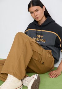 Brown cargo pants, gray and black hoodie with orange accents, beige high-top sneakers, seated on a green surface.