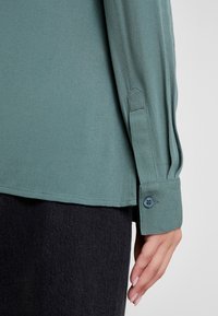 Ärmel und Manschette einer grünen Bluse mit langen Ärmeln und blauer Knöpfen, getragen über dunklen Hosen, mit einer entspannten Hand neben dem Körper.