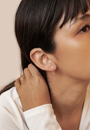 Goldene Ohrringe mit floralem Design und klaren Edelsteinakzenten, die an einer zarten Kette hängen und am Ohr eines Models getragen werden.